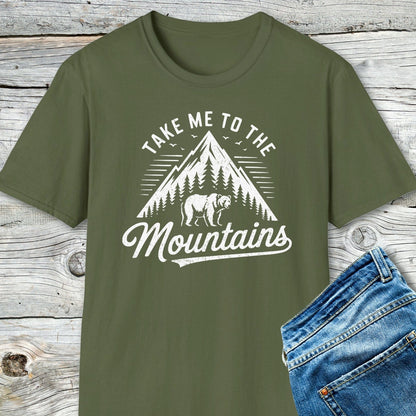 Take Me To The Mountains T-Shirt with mountain and bear motif, paired with blue jeans, showcased on a rustic wooden surface. Soft, lightweight cotton for adventure.