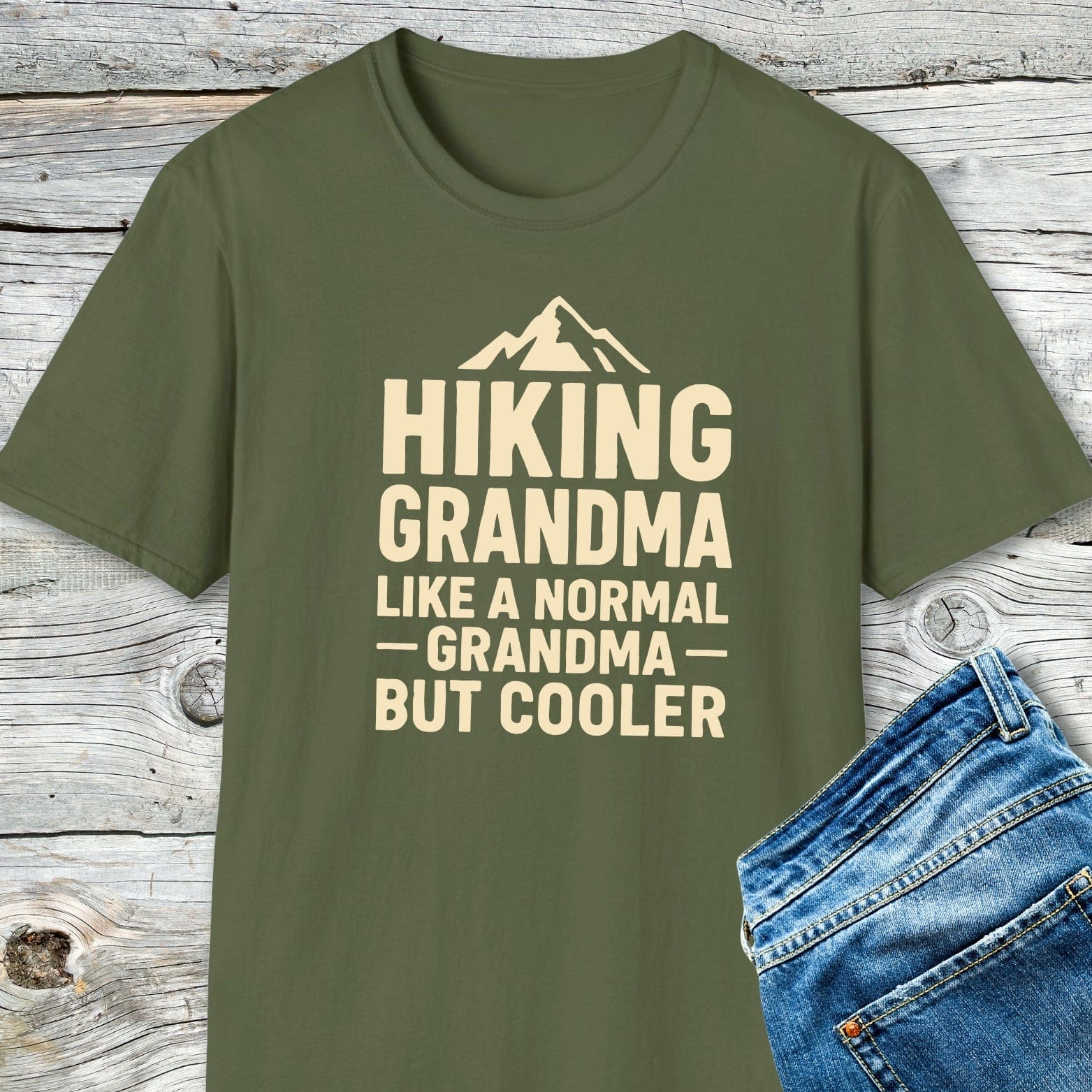 Hiking Grandma T-Shirt features a mountain graphic and bold text, paired with blue jeans on a wooden surface, emphasizing comfort and adventure-ready style.