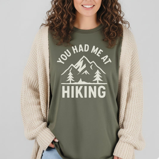 Person wearing You Had Me At Hiking T-Shirt with mountain graphic, under a cream cardigan, hands in light-wash jeans pockets, showcasing casual outdoor style.