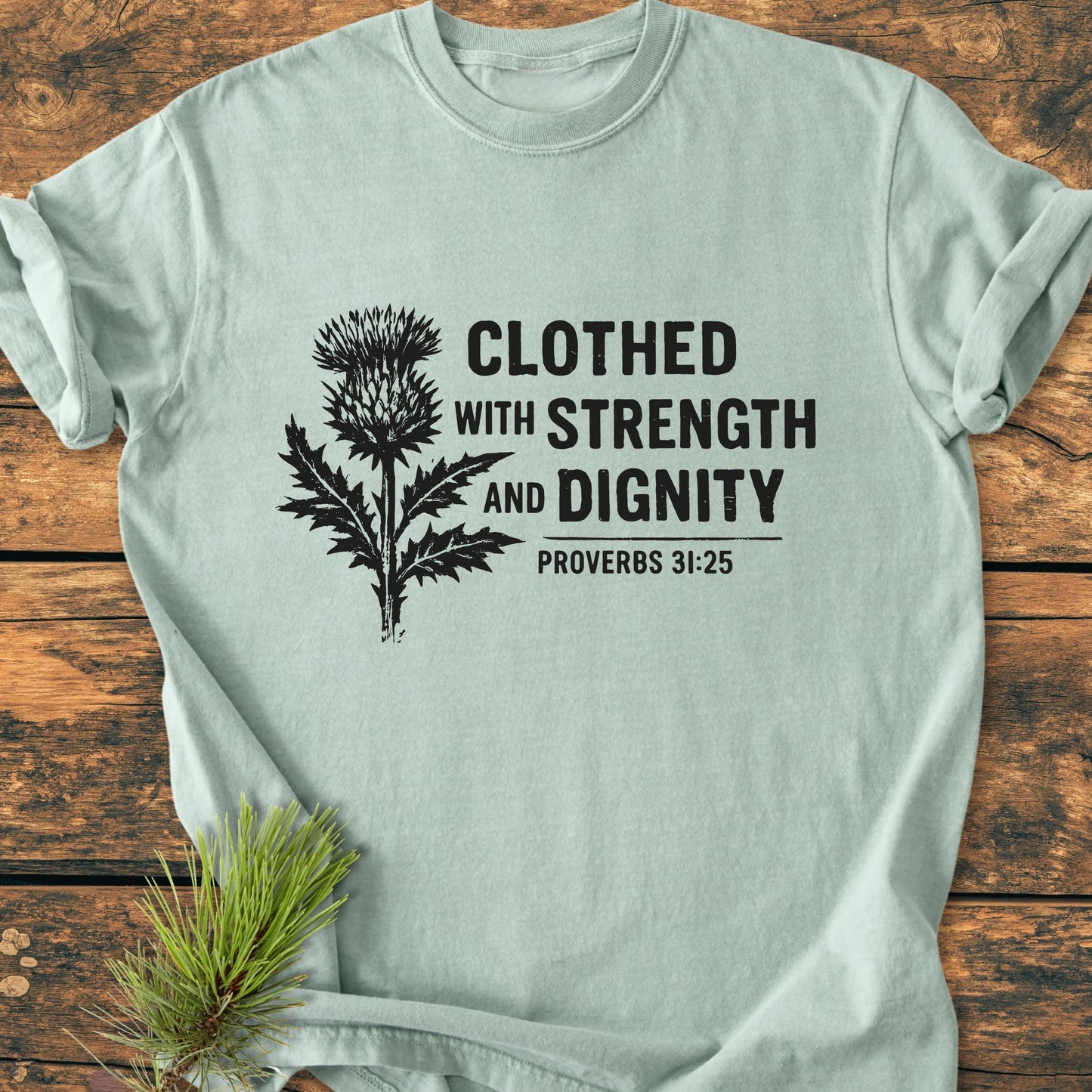 Soft-green tee featuring a black thistle illustration and Clothed With Strength And Dignity - Proverbs 31:25 text, laid flat with a pine sprig nearby, showcasing its relaxed, vintage wash design.