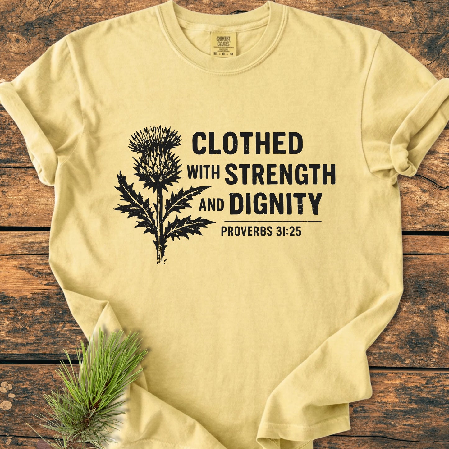 Clothed With Strength And Dignity - Vintage Wash Tee: Pale-yellow short-sleeve shirt with a black thistle illustration and Proverbs 31:25 text, laid flat with a pine branch accentuating its relaxed, vintage style.