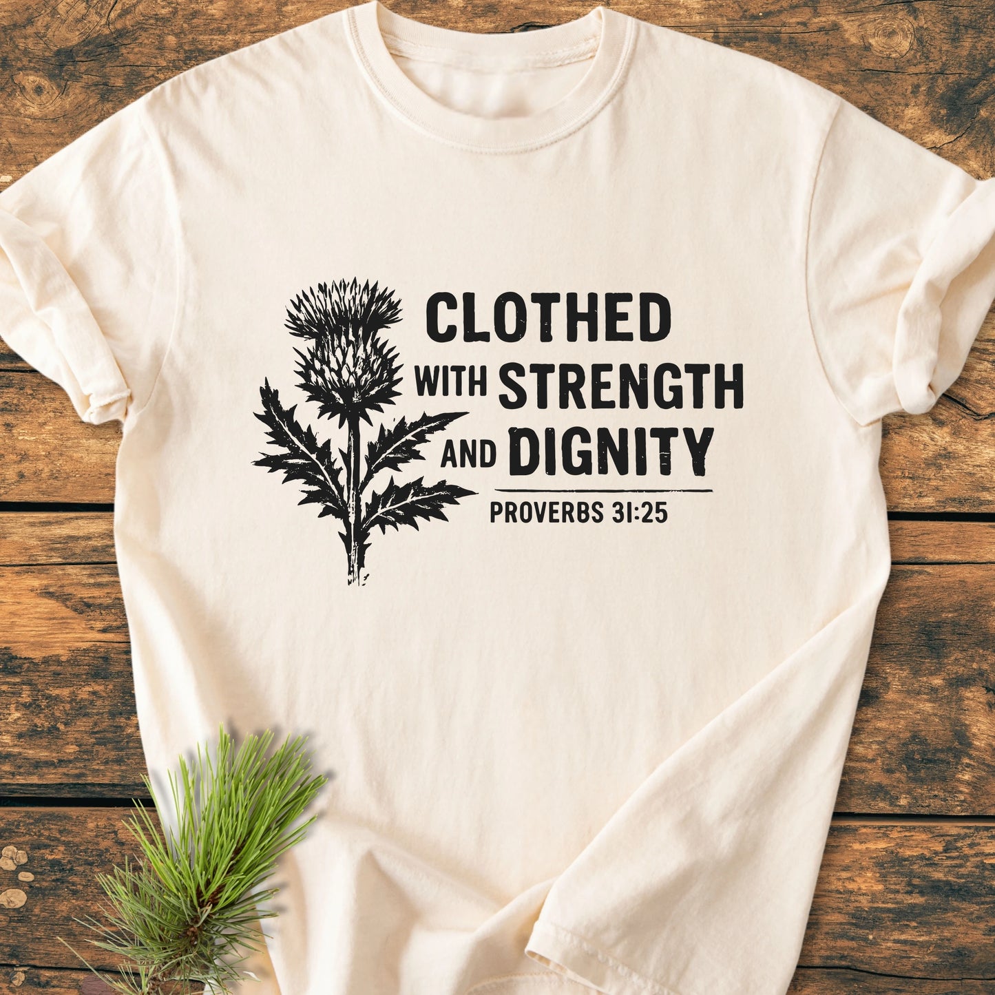 Light-cream T-shirt with a black stylized thistle and “CLOTHED WITH STRENGTH AND DIGNITY” on front, Proverbs 31:25 reference, showcasing the Clothed With Strength And Dignity - Vintage Wash Tee.