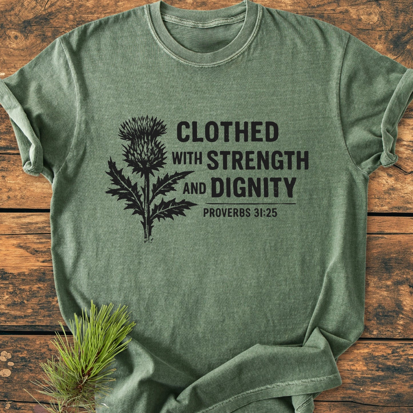 Clothed With Strength And Dignity - Vintage Wash Tee, featuring a black botanical print and Proverbs 31:25 text on soft olive-green fabric, laid on a wooden surface.