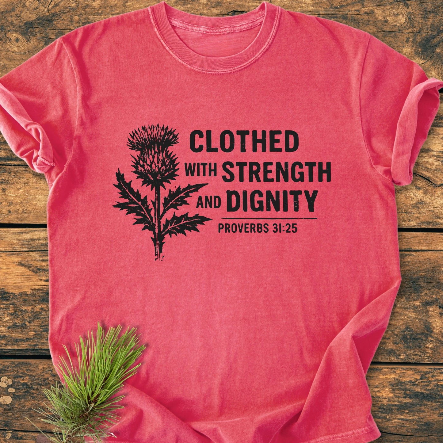 Clothed With Strength And Dignity - Vintage Wash Tee features a black thistle graphic and quote on a soft coral-pink shirt, laid flat with a pine sprig accent. Ideal for everyday wear.