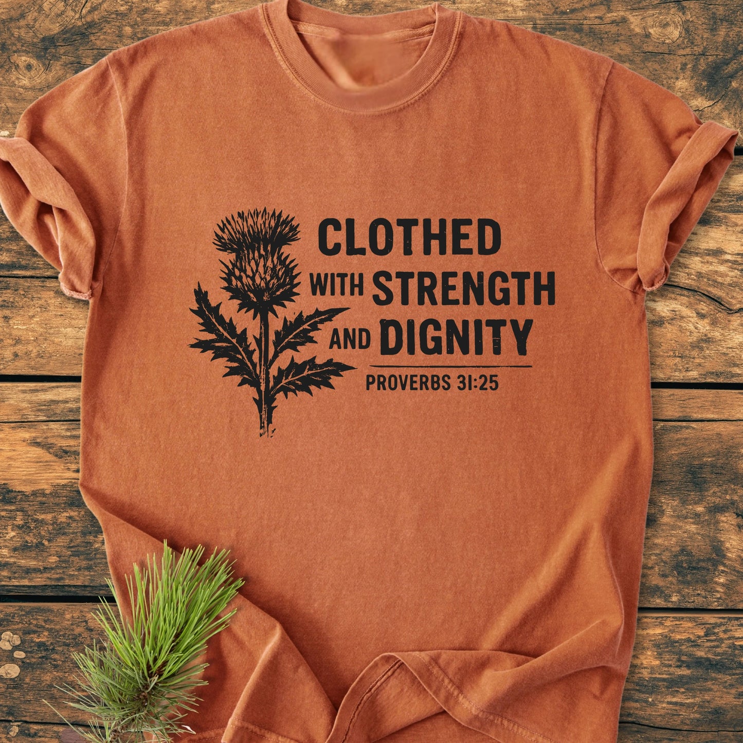 Clothed With Strength And Dignity - Vintage Wash Tee, featuring a black thistle design and quote, laid on wood with a pine sprig for styling, reflecting a relaxed, vintage-wash aesthetic.