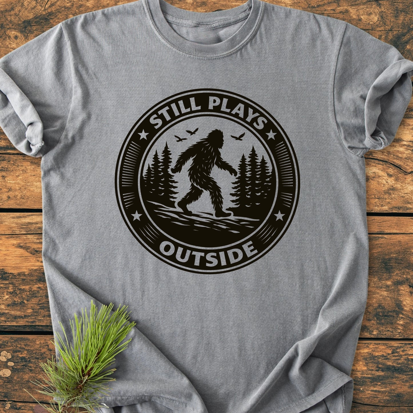 Gray 1430 Still Plays Outside Bigfoot T-shirt, featuring a black screen-printed badge with Bigfoot, pine trees, and birds, laid flat with an evergreen sprig, emphasizing its outdoor theme and vintage wash appeal.