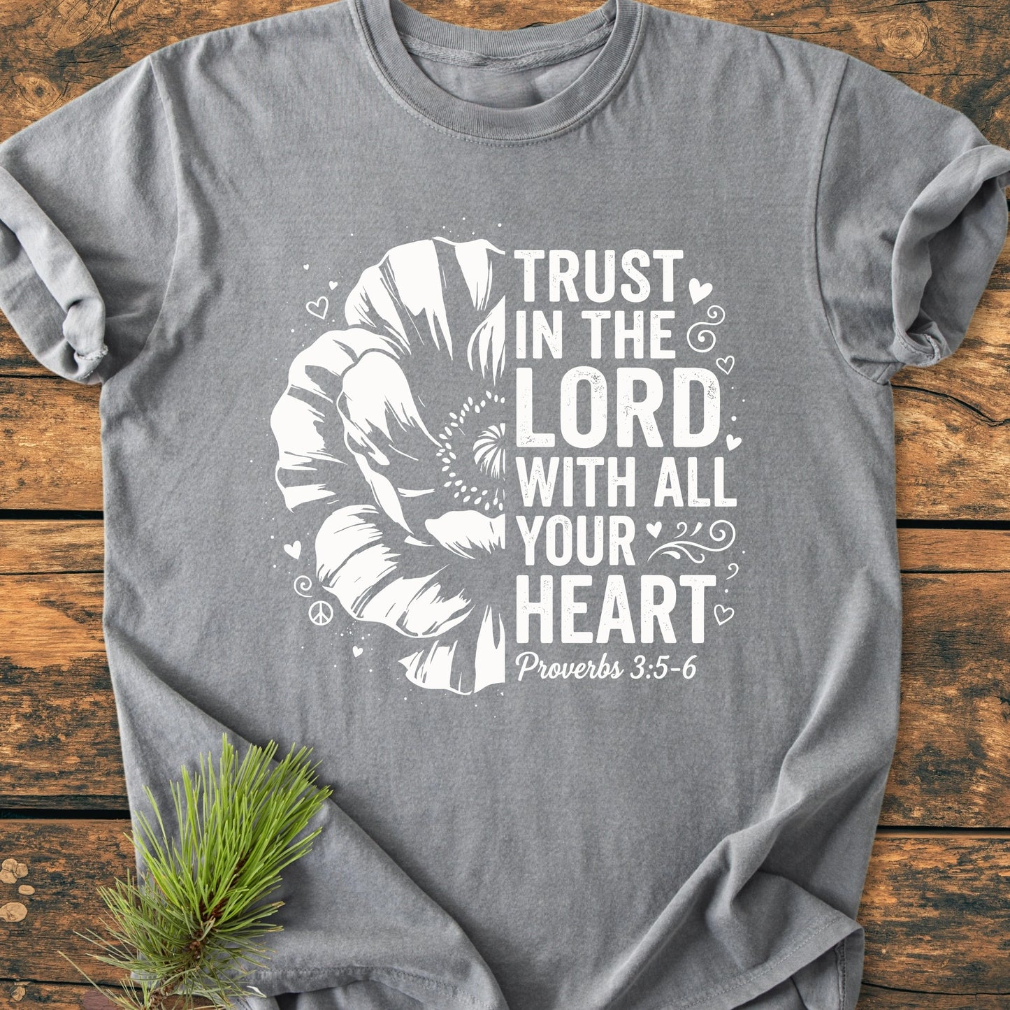 Trust In The Lord - Vintage Wash Tee features a stylized flower and Proverbs 3:5–6 text. Laid flat on wood, adorned with pine, showcasing relaxed fit and vintage character.