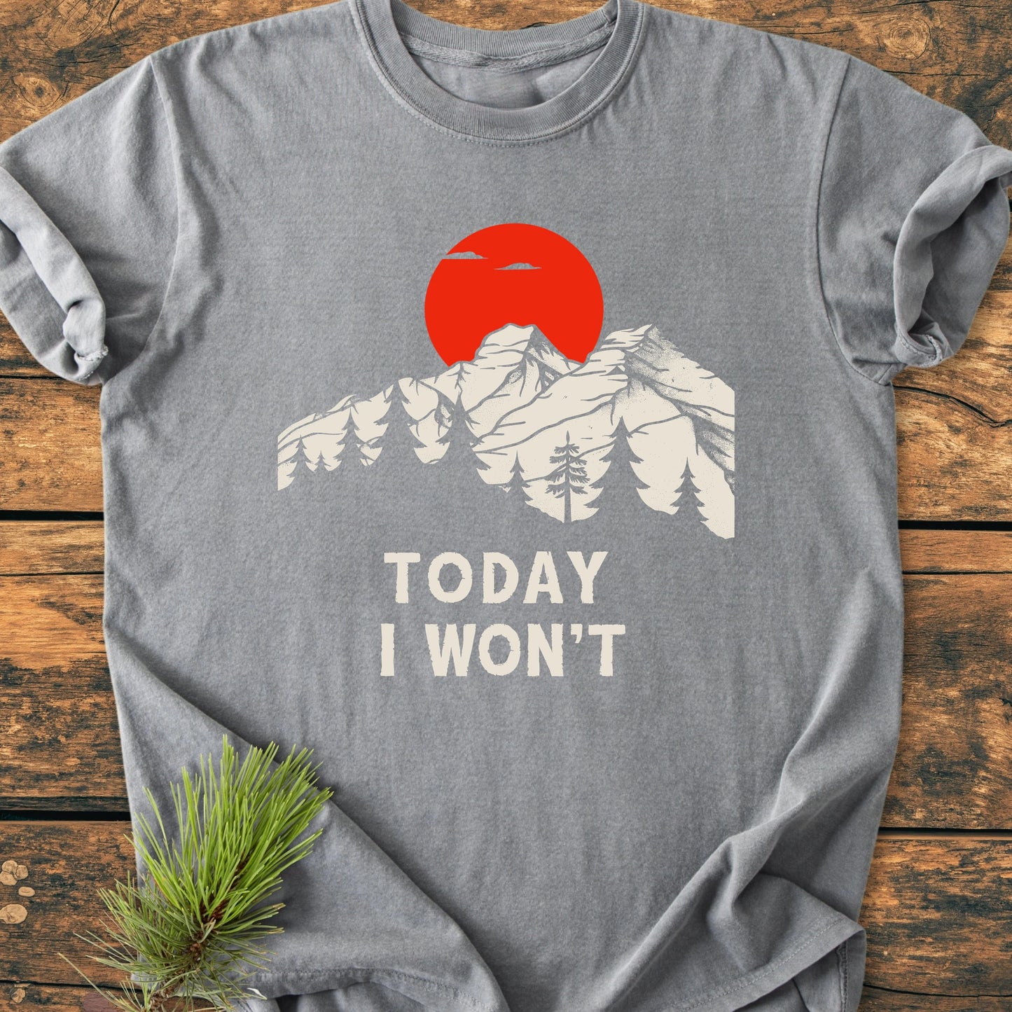 Heather-gray T-shirt featuring a stylized mountain range with pine trees and a bold red sun. Below, the phrase TODAY I WON'T in off-white. Sprig of pine needles tucked beneath.