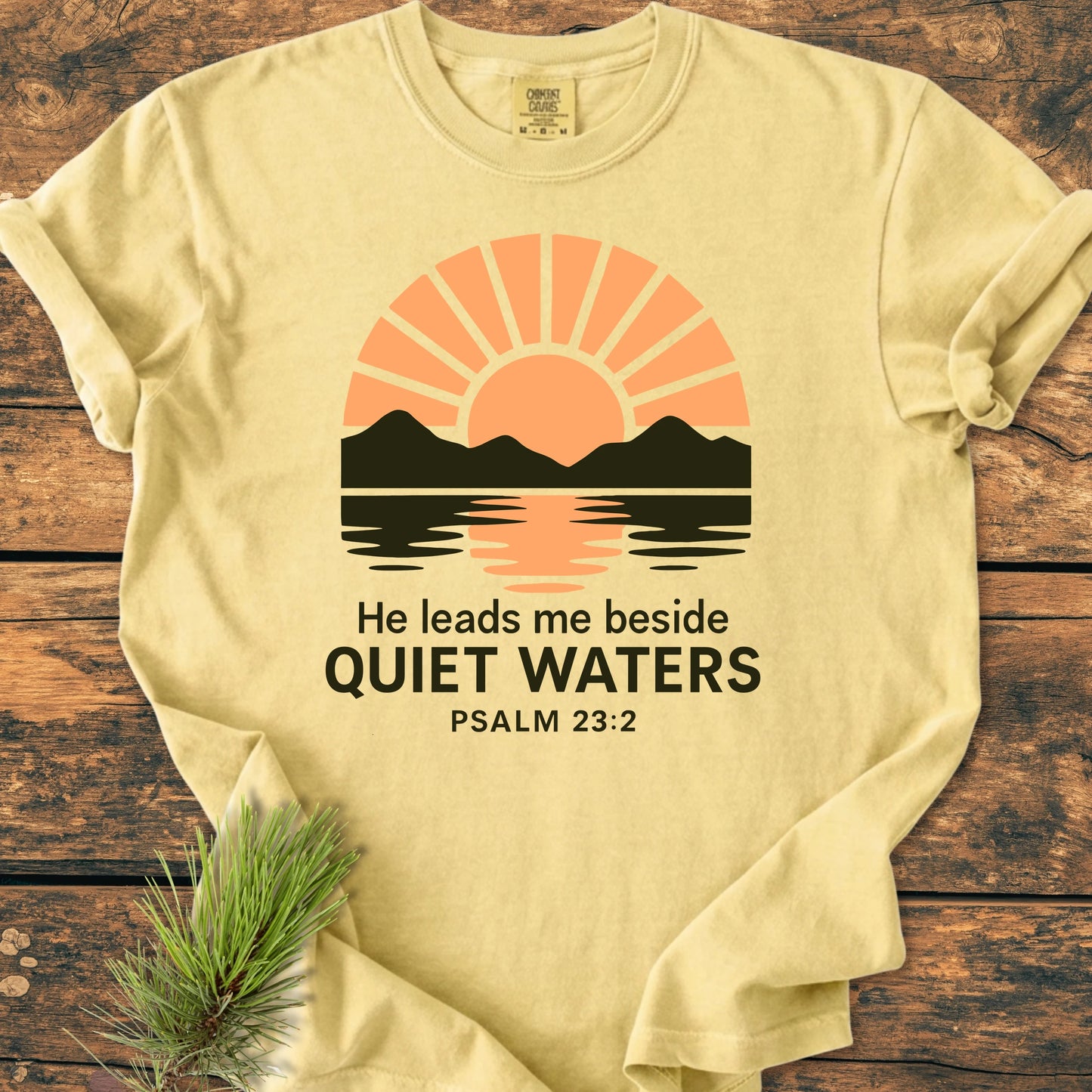 1424 He Leads Me T-shirt featuring a sun and mountain graphic with Psalm 23:2 text, displayed on a wooden surface with a pine sprig, emphasizing its vintage wash and relaxed fit.