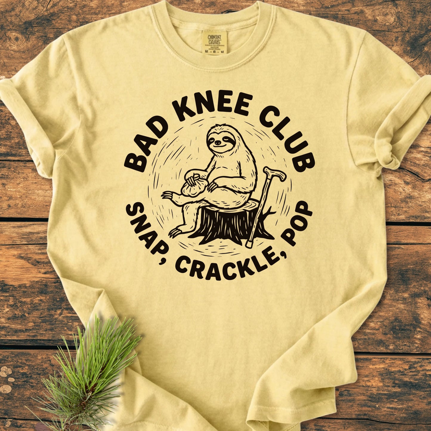 Pale-yellow T-shirt with BAD KNEE CLUB above a sloth illustration holding an ice-pack on a tree stump, cane beside it, and SNAP, CRACKLE, POP below, laid on wood surface.