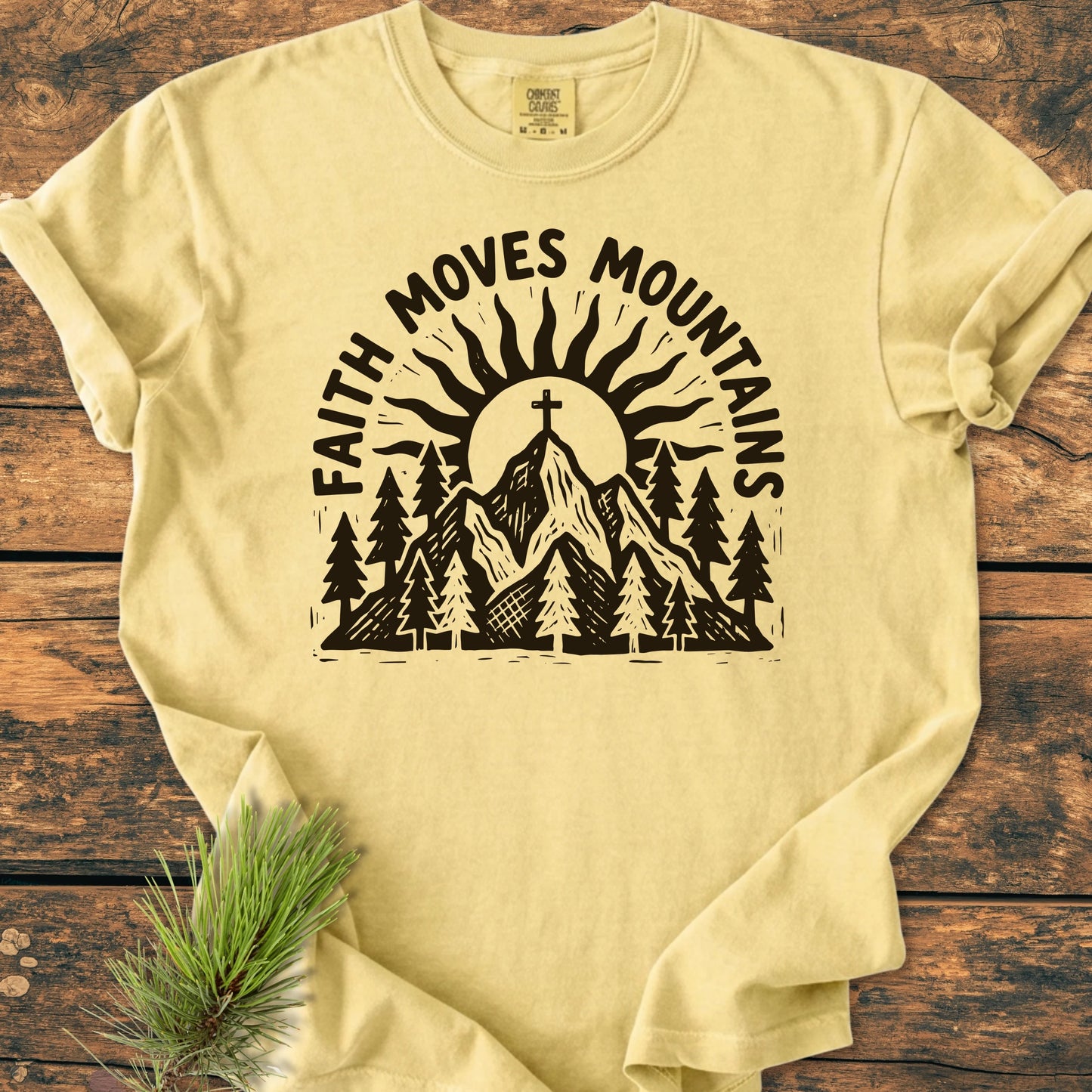 1409 Faith Moves Mountains T-shirt features a hand-drawn print of pine trees, mountains, and a sun, with a cross atop the tallest peak, on soft, garment-dyed fabric.