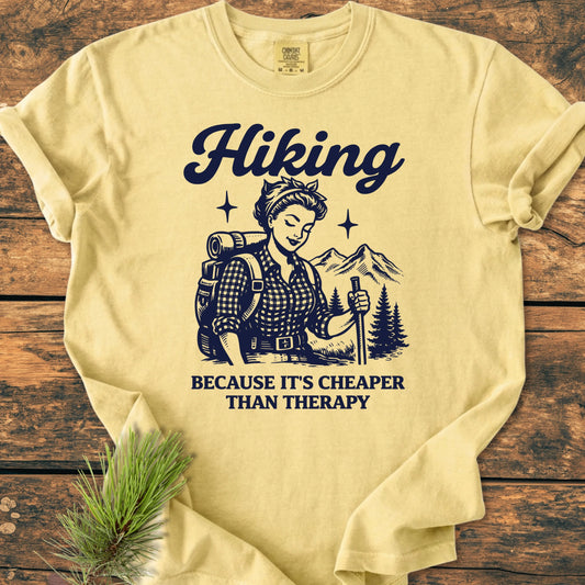 1407 Hiking Cheaper Than Therapy Base T-shirt: Pale-yellow tee featuring a vintage-style illustration of a woman hiker with pine trees and mountains, emphasizing outdoor adventure and relaxation.