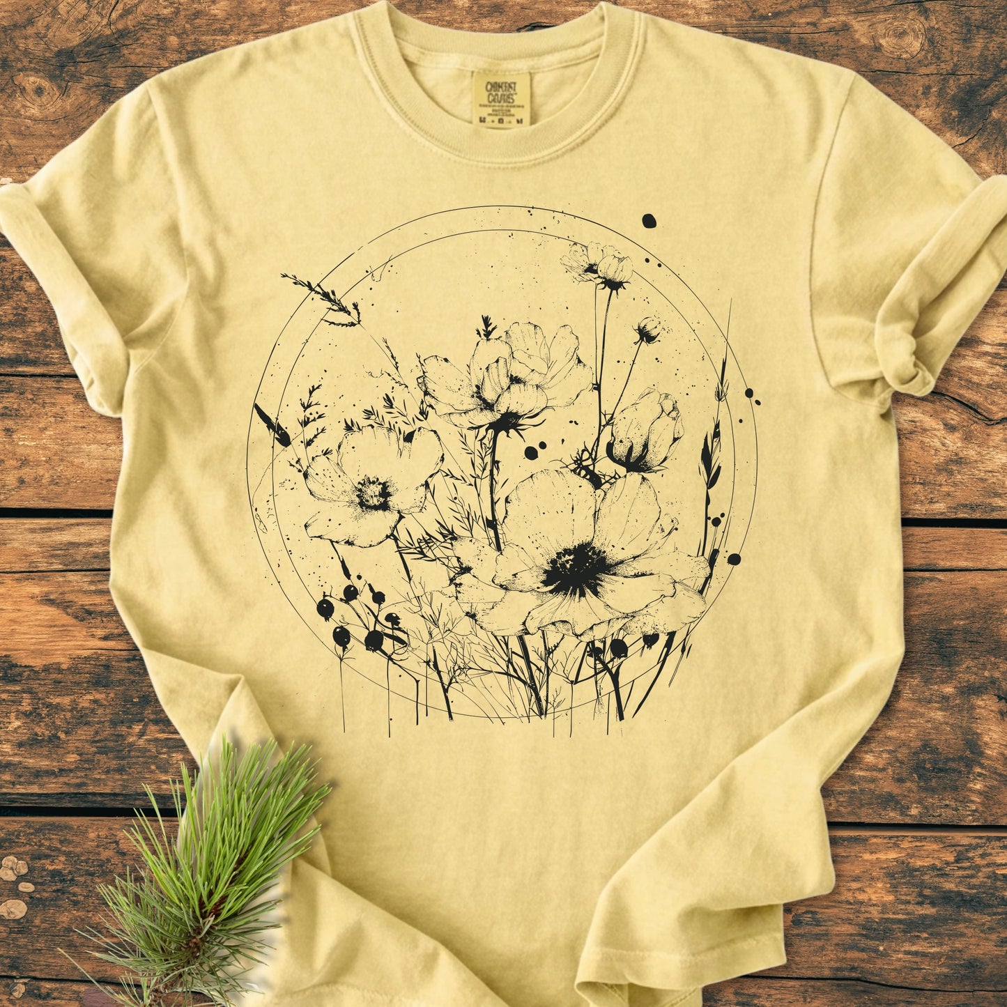 391 Wildflower Circle T-shirt features a black wildflower illustration in a circle on a pale yellow surface, laid on wood. Pine needles accentuate the natural, rustic appeal.