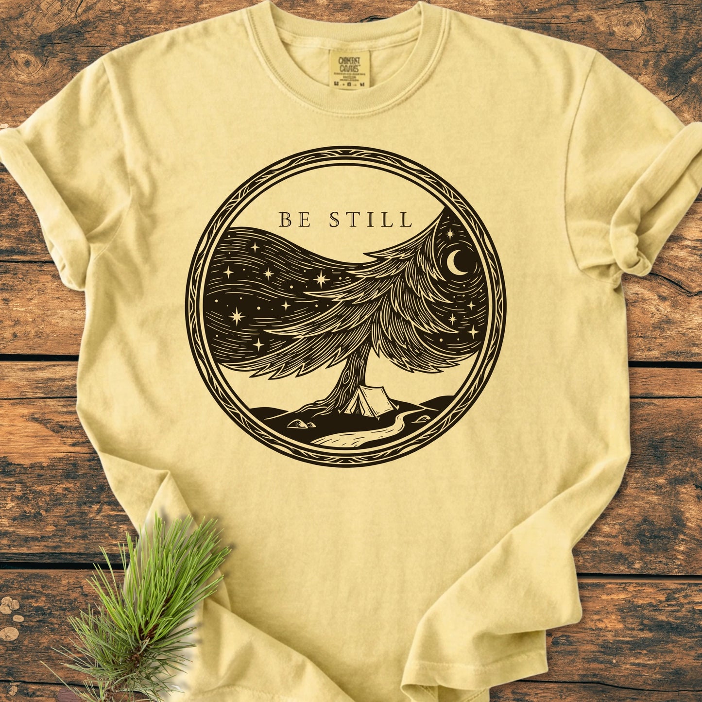 1411 Be Still T-shirt features a woodcut-style graphic of a windswept tree, starry sky, and crescent moon, highlighting the words BE STILL in uppercase above.
