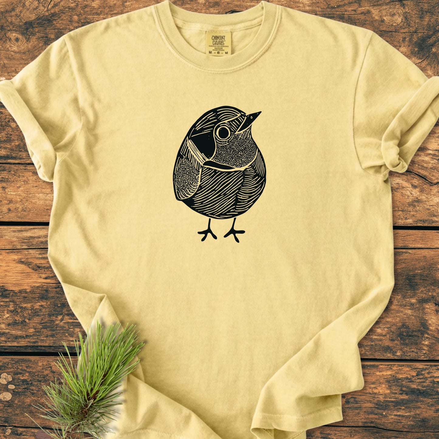 Cream T-shirt titled 1433 Little Robin featuring a black line-drawing of a bird, laid flat on wooden surface with a pine sprig, embodying a relaxed, vintage-wash style.