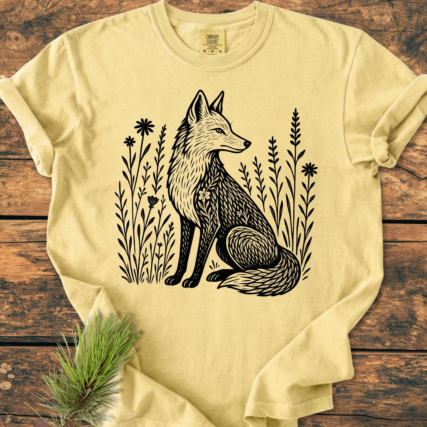 225 Wildflower Fox DARK T-shirt features a black fox illustration amid wildflowers on a pale-yellow tee, laid flat on wood. Includes a pine sprig accent. Made from soft, durable 100% ring-spun cotton.
