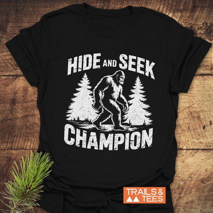 Hide And Seek Champion T-Shirt featuring a black and white illustration of Bigfoot walking, accompanied by white text on the shirt.