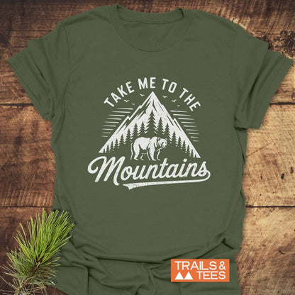 Take Me To The Mountains T-Shirt featuring a mountain, trees, and bear graphic. Includes “TRAILS & TEES” badge; laid on wood with pine needles.