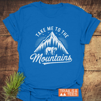 Take Me To The Mountains T-Shirt featuring a mountain and bear graphic, styled on wood with a pine branch, showcasing outdoor adventure appeal.