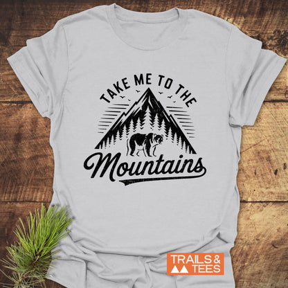 Take Me To The Mountains T-Shirt featuring a mountain, trees, and bear graphic; paired with a pine sprig, highlighting outdoor adventure.