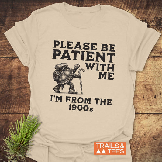 Please Be Patient With Me I'm From The 1900s T-Shirt featuring a cartoon hiking tortoise, brand logo visible, laid on a wooden surface.