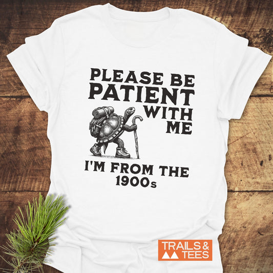 Please Be Patient With Me I'm From The 1900s T-Shirt features a turtle illustration with hiking gear, lying on a wooden surface with a pine sprig beside it.
