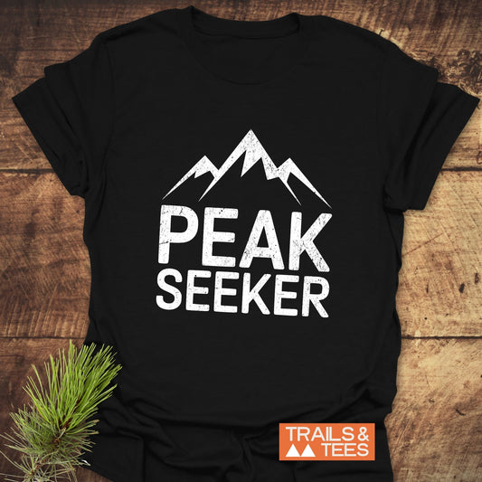 Peak Seeker T-Shirt featuring three mountain peaks graphic, “PEAK SEEKER” text, styled with pine sprig, embodies adventure with soft, durable, ring-spun cotton.
