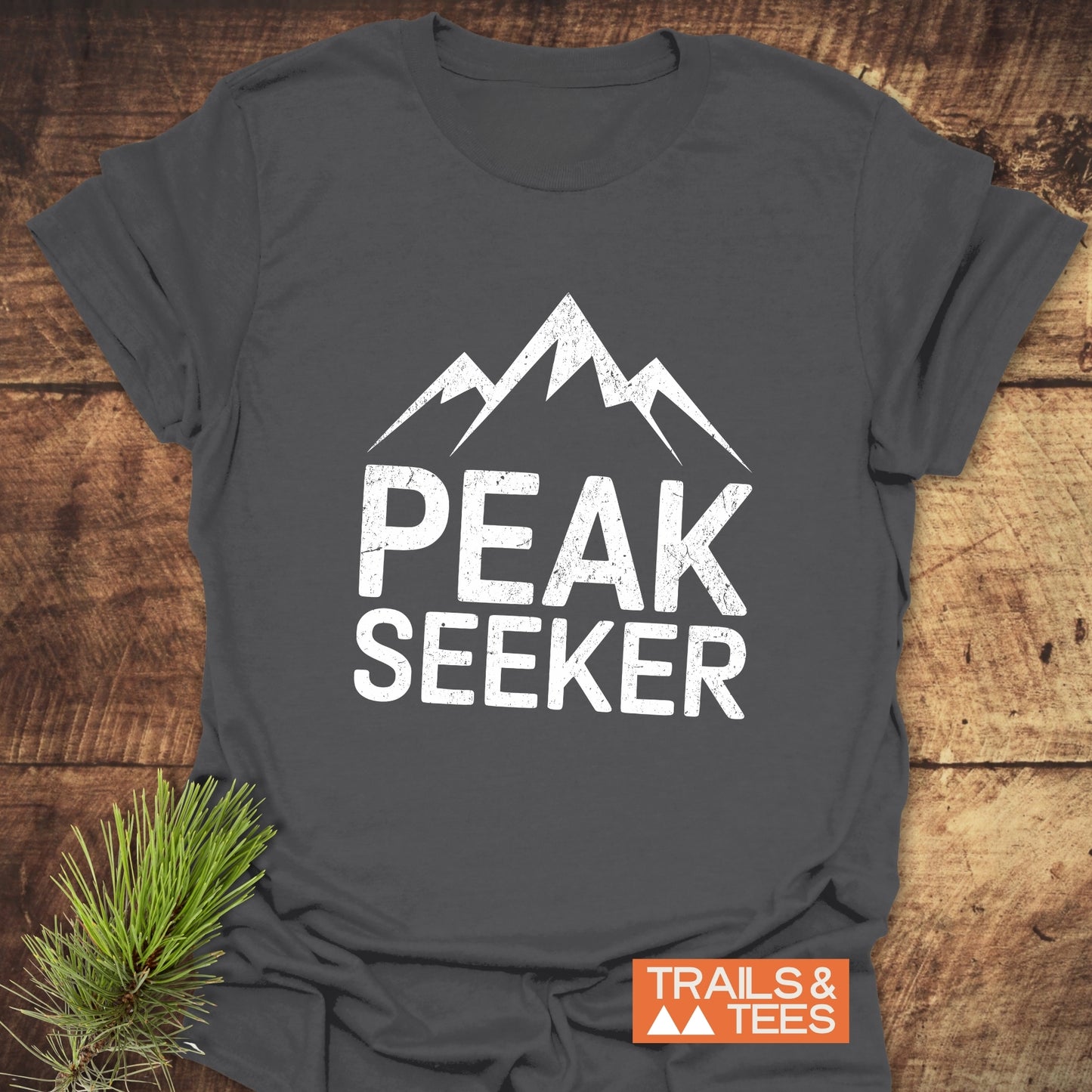 Charcoal-gray Peak Seeker T-shirt featuring a mountain graphic, laid on wood. Soft, ring-spun cotton for outdoor adventures. Includes TRAILS & TEES logo.