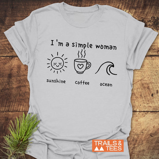 I'm A Simple Woman - Sunshine Coffee Ocean T-Shirt on a wooden surface, featuring black line drawings. Includes TRAILS & TEES logo and a pine sprig beside it.