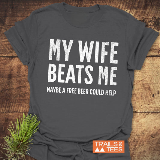 Dark gray My Wife Beats Me - Free Beer T-shirt with distressed white text, orange TRAILS & TEES logo, styled with pine sprig, on wooden surface.