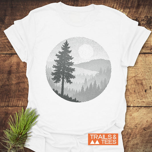 Sunset Woods Halftone T-Shirt with a black halftone landscape graphic, showcasing pine tree, hills, and sun, on soft, ring-spun cotton, laid on wooden surface.
