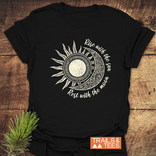 Sun & Moon Mystical Boho T-Shirt featuring a half-sun/half-moon graphic with the motto Rise with the sun, Rest with the moon, laid on wood.