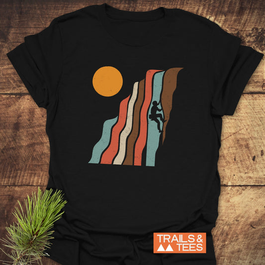 Retro Climber T-Shirt featuring a rock climber graphic with wavy stripes and sun, displayed on wood. Includes TRAILS & TEES label, pine sprig nearby.
