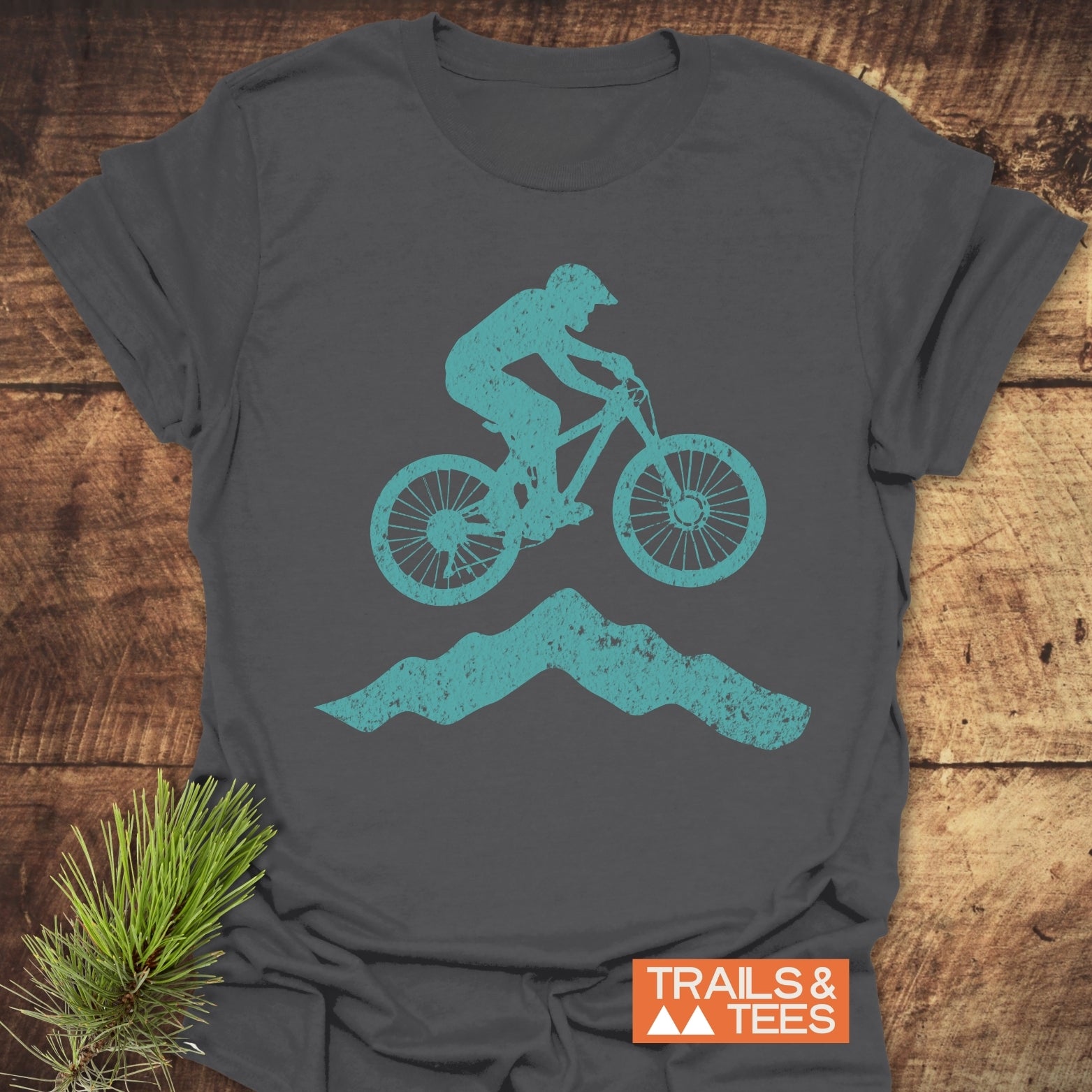 Airborne Mountainbiker T-Shirt featuring a teal biker graphic on charcoal-gray cotton, laid flat with a TRAILS & TEES logo. Perfect for outdoor adventures.