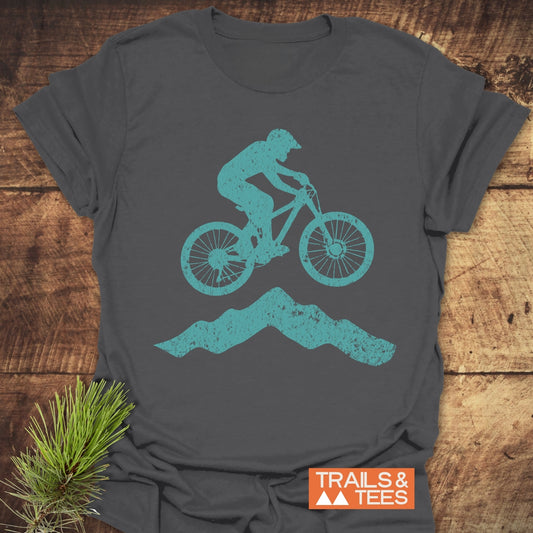 Airborne Mountainbiker T-Shirt featuring a teal biker graphic on charcoal-gray cotton, laid flat with a TRAILS & TEES logo. Perfect for outdoor adventures.