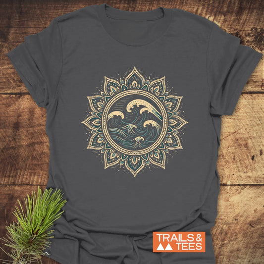 Ocean Mandala T-Shirt on wooden surface, featuring cream and teal ocean wave graphic within lotus mandala pattern. Includes “TRAILS & TEES” logo and pine sprig accent.