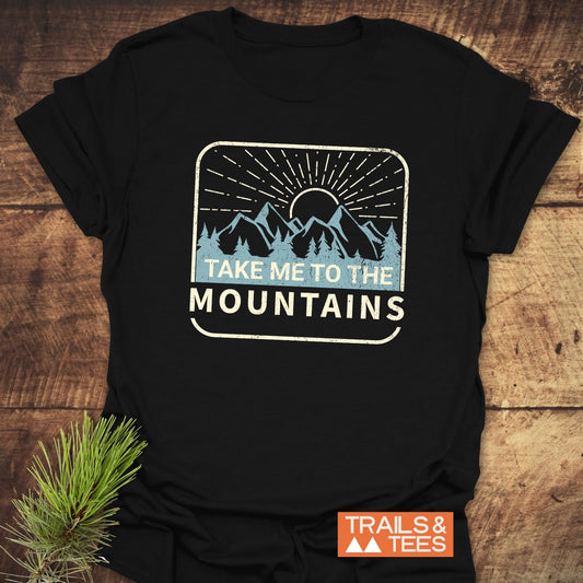 Take Me To The Mountains T-Shirt with mountain graphic on black fabric, laid on wood. Features durable, comfortable ring-spun cotton for outdoor adventures.