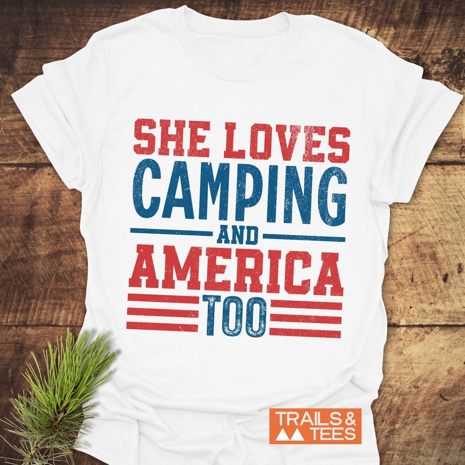 She Loves Camping T-Shirt on wood surface, featuring distressed red and blue text. Includes TRAILS & TEES. logo. Made from soft, lightweight cotton for outdoor adventures.