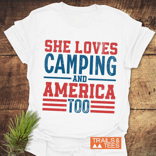 She Loves Camping T-Shirt on wood surface, featuring distressed red and blue text. Includes TRAILS & TEES. logo. Made from soft, lightweight cotton for outdoor adventures.