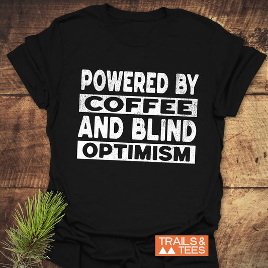 Black T-shirt with POWERED BY COFFEE AND BLIND OPTIMISM text; features durable print, soft ring-spun cotton, and TRAILS & TEES logo, ideal for outdoor enthusiasts.