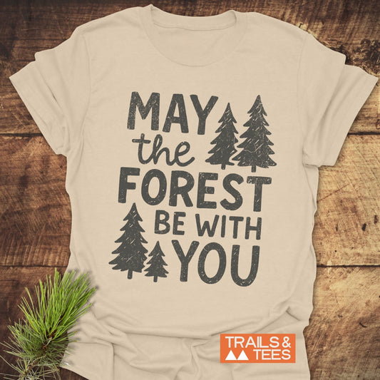 May The Forest Be With You T-Shirt featuring distressed print and evergreen motifs, laid on wood with a pine sprig, embodying an outdoor adventure vibe.