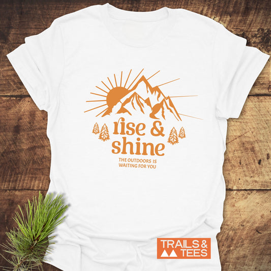 Rise And Shine T-Shirt with orange sun and mountains graphic, soft ring-spun cotton, designed for outdoor enthusiasts, featuring durable prints and a TRAILS & TEES logo.