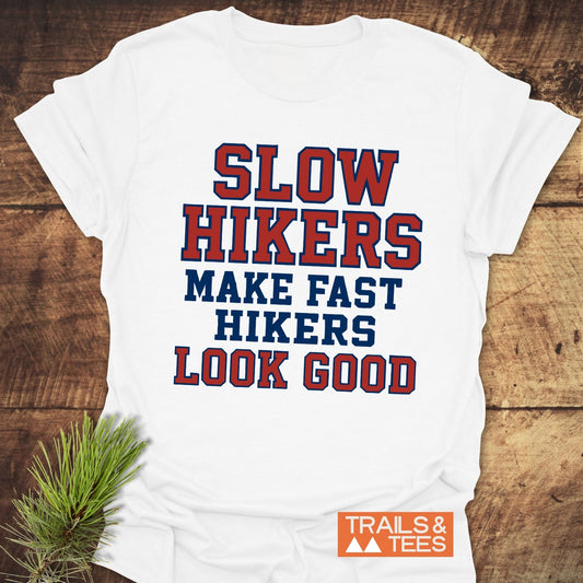 “Slow Hikers Make Fast Hikers Look Good” T-Shirt on wood, featuring bold slogan in red and blue. Made from soft, lightweight ring-spun cotton for comfort.
