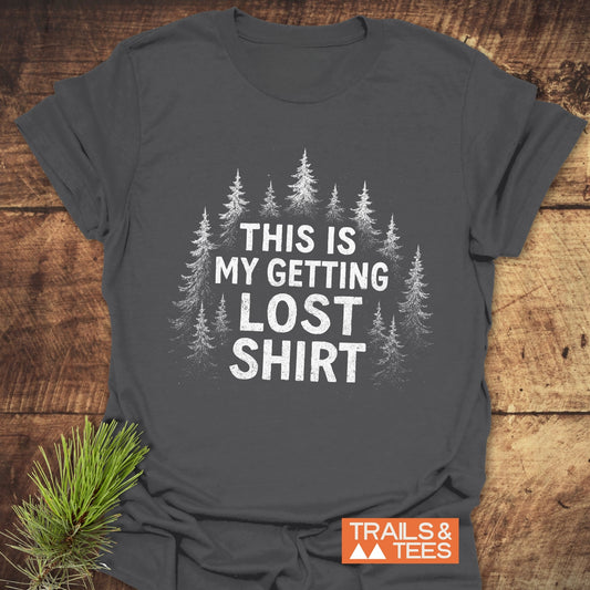 Getting Lost T-Shirt with pine tree design and THIS IS MY GETTING LOST SHIRT text, featuring Trails & Tees logo, laid on wooden surface with pine sprig.