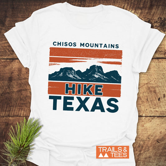Chisos Mountains Texas T-Shirt with a mountain graphic, crafted from soft cotton for outdoor enthusiasts, laid on wood with evergreen sprig and Trails & Tees logo.