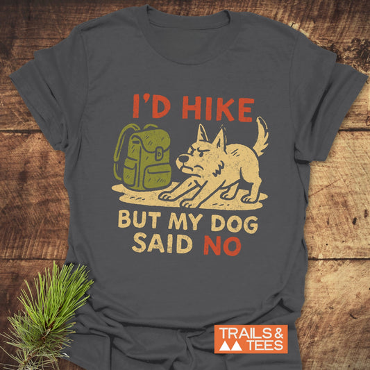 Dog Stops The Hike T-Shirt featuring a stubborn dog graphic and humorous text, crafted from soft cotton for adventure-ready comfort, beside a pine sprig on wood.