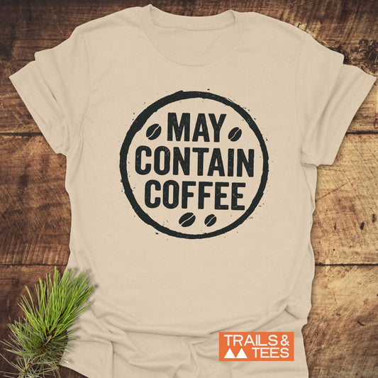 May Contain Coffee T-Shirt with a round graphic, crafted from soft ring-spun cotton, laid on a wooden surface with a pine sprig and Trails & Tees label.