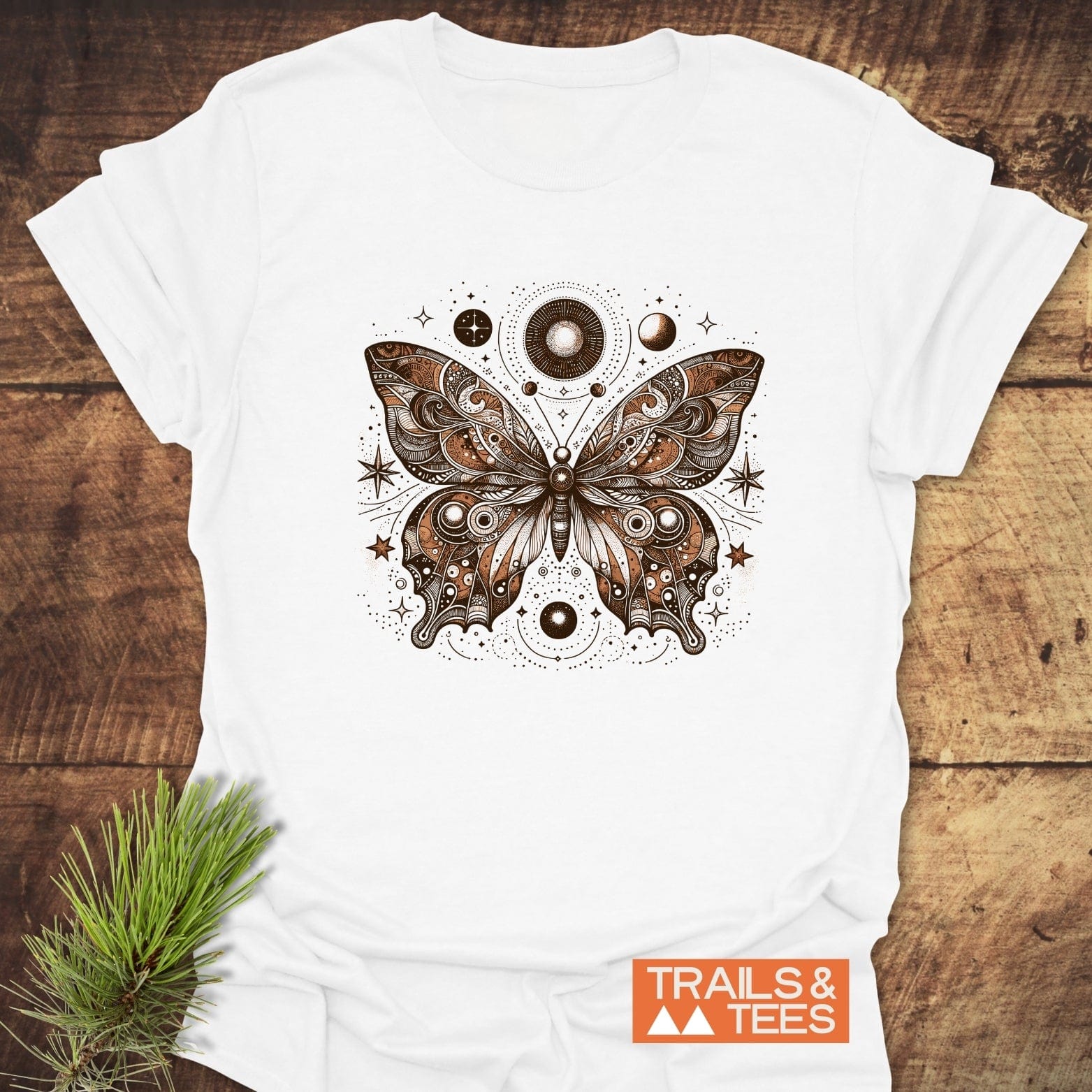 Celestial Butterfly T-Shirt featuring a detailed butterfly with celestial motifs on soft cotton, laid on wood beside a pine sprig, with TRAILS & TEES logo.