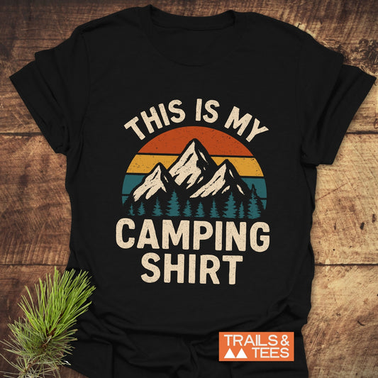 Black T-shirt with retro mountain graphic and “THIS IS MY CAMPING SHIRT” text, showcasing an adventure-ready design by Trails & Tees, laid on wood.