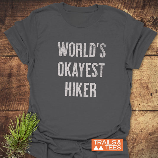 World's Okayest Hiker T-Shirt on wood, featuring distressed white text and Trails & Tees logo, with pine sprig accentuating outdoor theme.